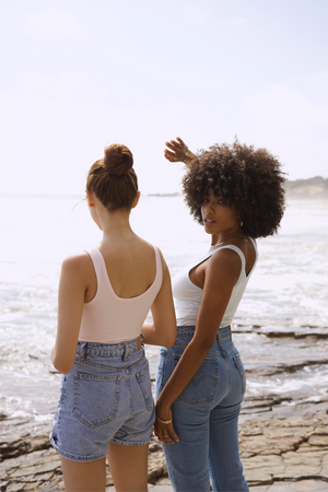 Two women wearing Ambitionist Everyday Classic Tank Bodysuits in baby pink and white with denim on the beach